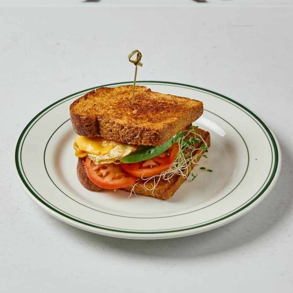 A breakfast sandwich with fried egg, cheese, tomato, avocado, and sprouts on toasted bread.