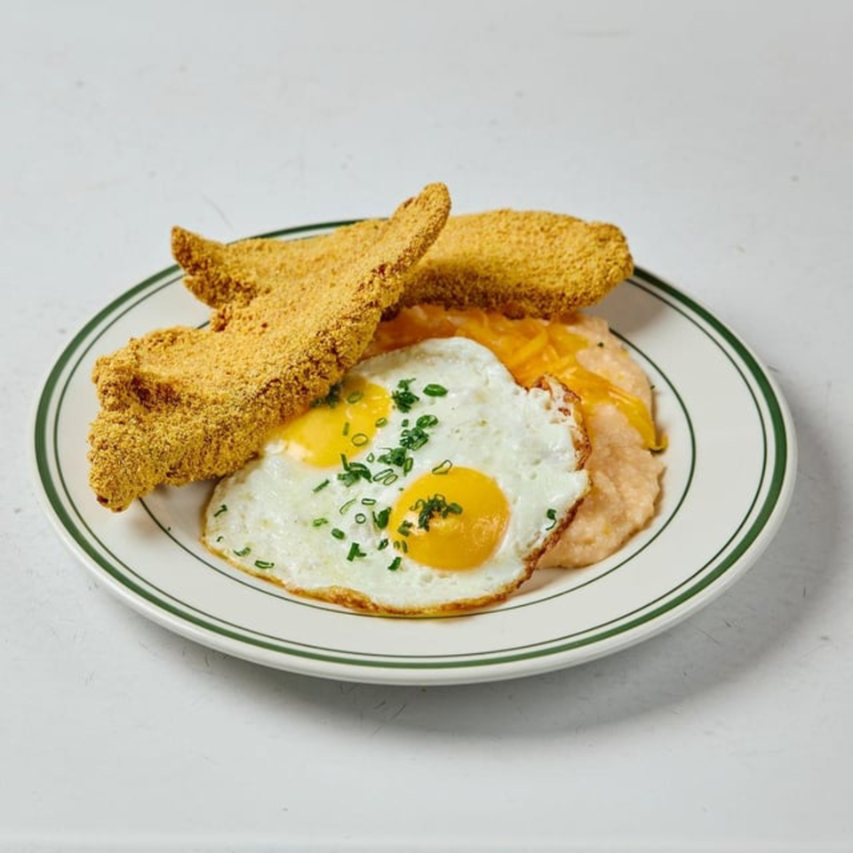 Fried catfish, two sunny-side-up eggs with chives, and cheesy grits on a white plate.