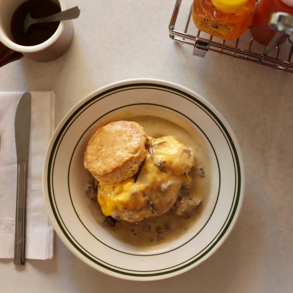 A bowl of biscuits and gravy with melted cheese, a coffee cup, and honey bottles.